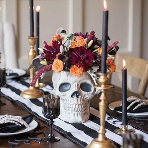 a stylish modern Halloween table setting with bold blooms in a skull, deep toned candles, black and white linens and skeleton hands
