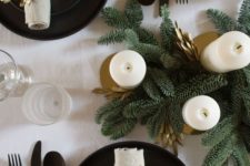a stylish winter tablescape with evergreens, pillar candles, black plates and chargers and cutlery plus gold branches