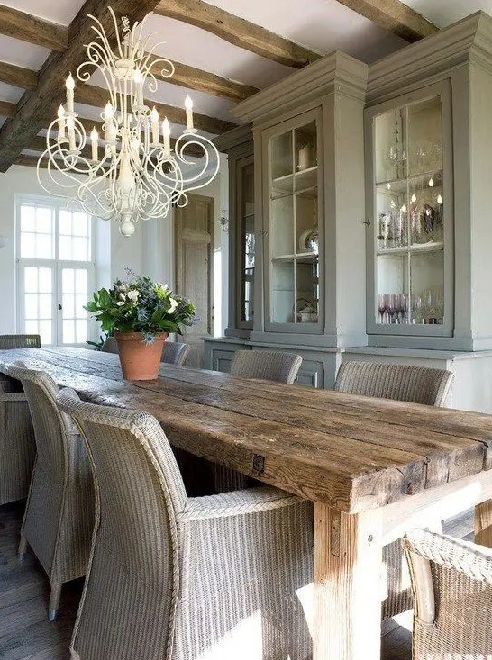 a vintage meets rustic dining room with stained wooden beams on the ceiling, a light grey storage unit, a stained wooden table and wicker chairs, a chic creamy chandelier