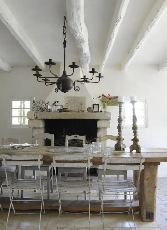 a vintage rustic dining room done in neutrals, with a large hearth, a wooden dining table and chairs, a black metal chandelier and lots of greenery