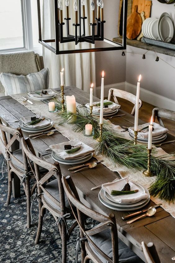 a winter table setting with an evergreen table runner, various candles, bells, neutral porcelain