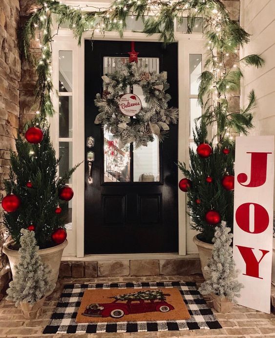 a bright holiday porch with mini trees with red ornaments, mini frosted trees, a frozen wreath and an evergreen garland with lights