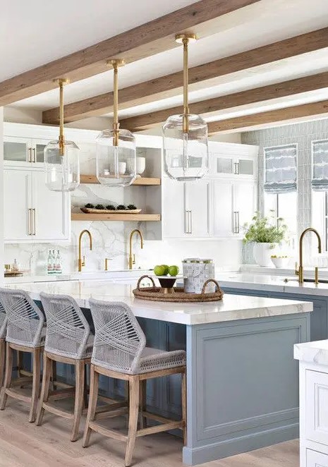 a fab coastal kitchen with white shaker style cabinets and a light blue kitchen island, glass pendant lamps and grey woven stools plus gold fixtures