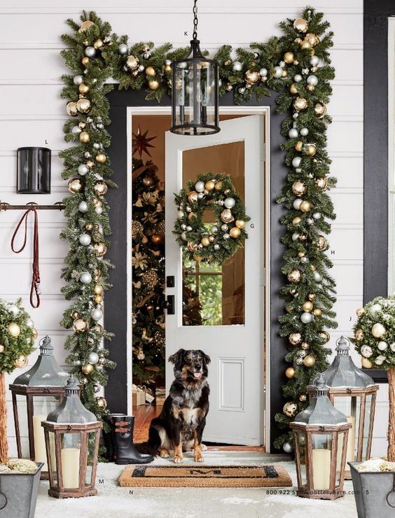 a festive Christmas porch with wooden candle lanterns, an evergreen garland with metallic ornaments and a matching wreath on the door