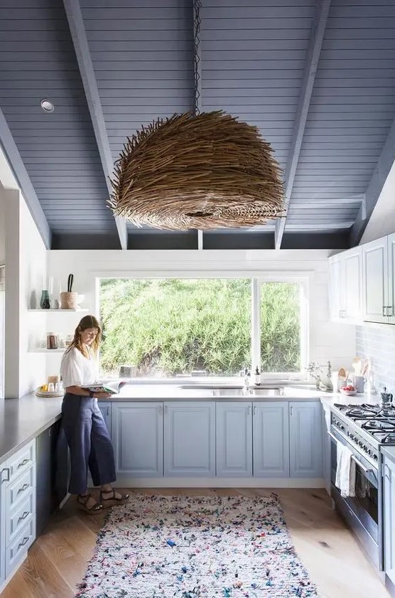 a lovely beach kitchen with light blue cabinets, a window instead of a backsplash, a unique woven pendant lamp