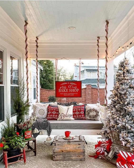 a lovely farmhouse Christmas porch with a hanging bench, mini and usual Christmas trees, red touches and lights is wow