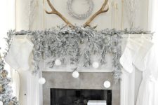 a refined Christmas mantel with a sivler evergreen garland, white ornaments, antlers, white stockings and white gift boxes