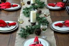 a rustic Christmas table with a burlap runner, metal chargers, red napkins, an evergreen runner, pinecones and candles