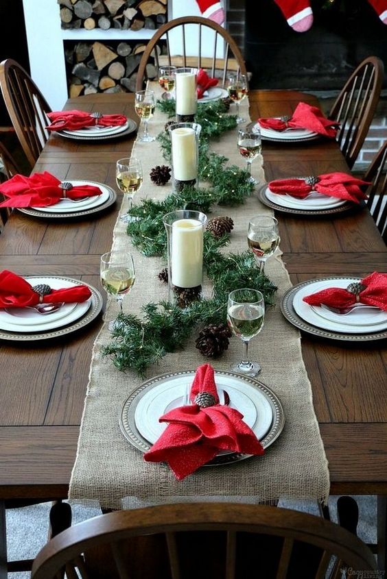 a rustic Christmas table with a burlap runner, metal chargers, red napkins, an evergreen runner, pinecones and candles