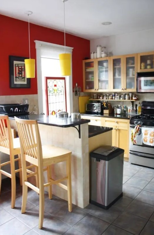 a small bright kitchen with a statement red wall, yellow pendant lamps, light stained furniture and a tall countertop for eating