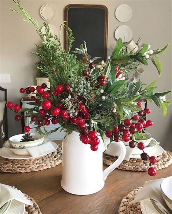 a white jug with lots of textural greenery, berries and fruits, woven placemats and printed porcelain for a rustic Christmas