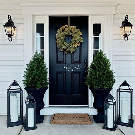 an elegant Christmas porch with a foliage wreath, Christmas trees in black urns, candle lanterns and a rug at the door