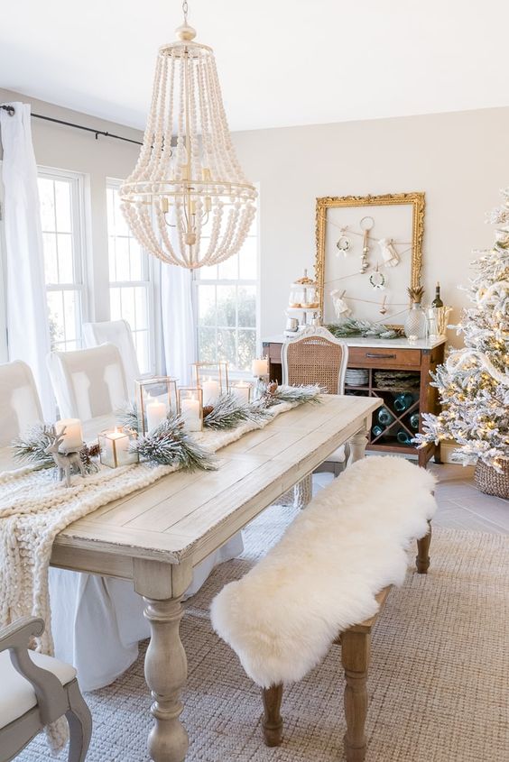 an exquisite white vintage Christmas space with a white table and chairs, a white knit table runner, white faux fur, flocked evergreens and candles and a flocked Christmas tree