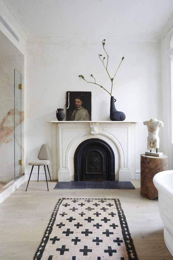 a Parisian bathroom with a vintage non-working fireplace, a glass-enclosed shower space, a bathtub, a printed rug, refined and chic artworks