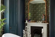 a Victorian style bathroom with moody wallpaper walls, a non-working fireplace, a clawfoot bathtub, navy wardrobes, a crystal chandelier and plants