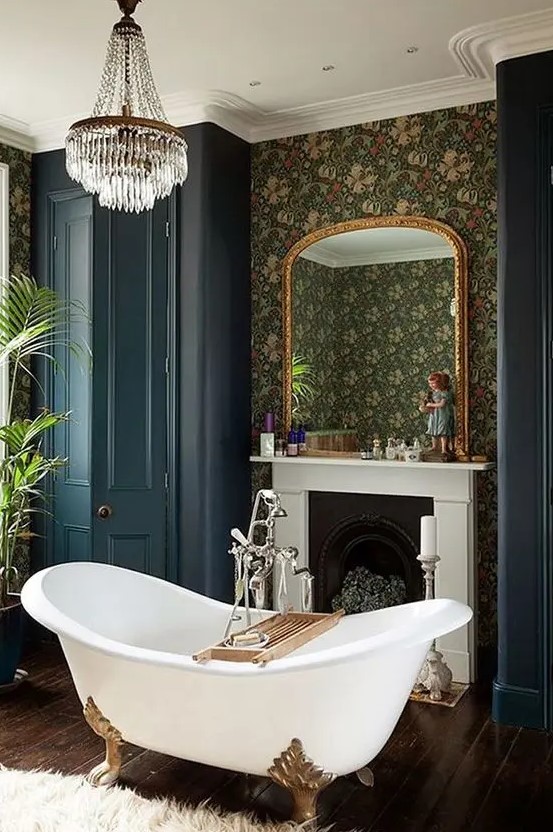 a Victorian style bathroom with moody wallpaper walls, a non working fireplace, a clawfoot bathtub, navy wardrobes, a crystal chandelier and plants