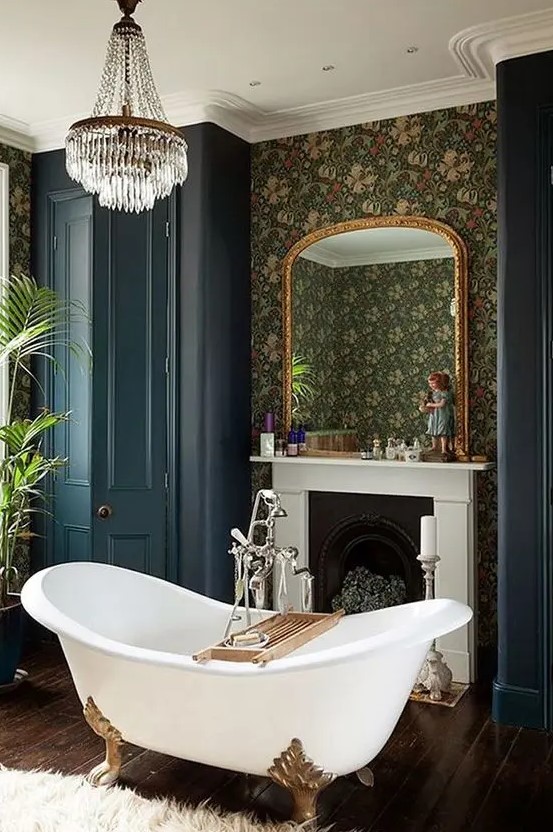 a Victorian style bathroom with moody wallpaper walls, a non-working fireplace, a clawfoot bathtub, navy wardrobes, a crystal chandelier and plants