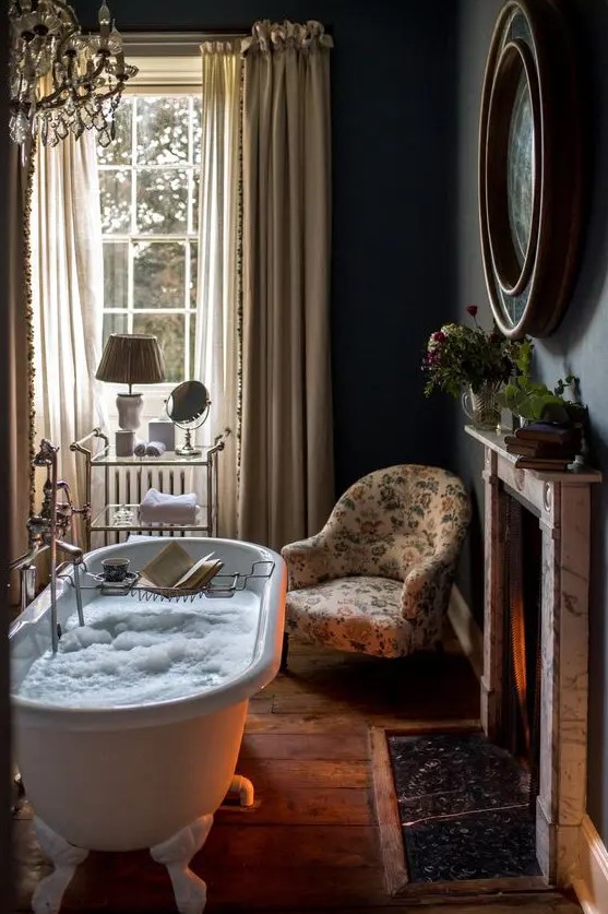 a beautiful vintage bathroom with black walls, a fireplace, a clawfoot bathtub, neutral curtains, a floral chair and some greenery and blooms