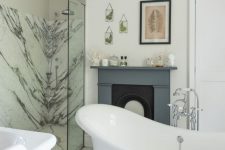 a catchy bathroom with white marble tiles, a white marble shower, a free-standing bathtub, a fireplace with a grey mantel and leaf art