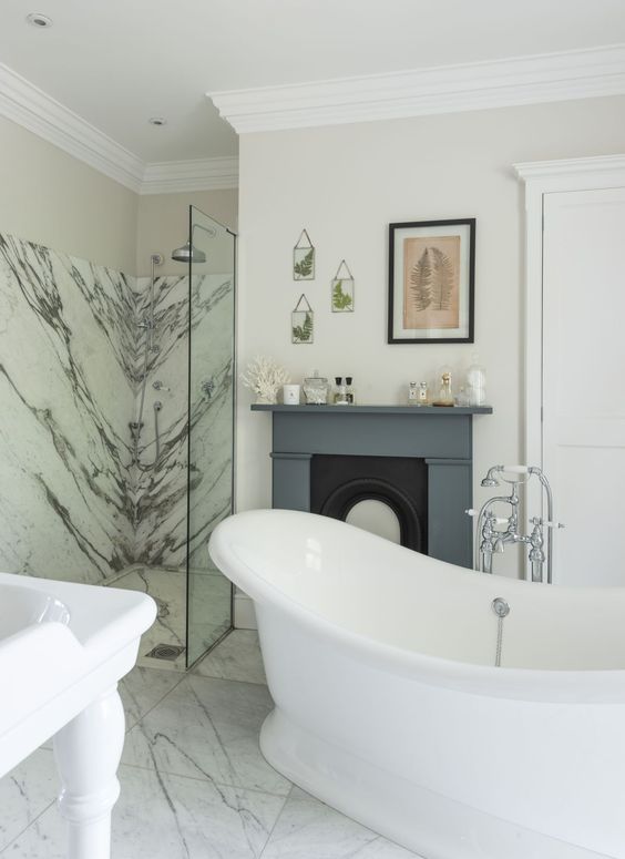 a catchy bathroom with white marble tiles, a white marble shower, a free standing bathtub, a fireplace with a grey mantel and leaf art