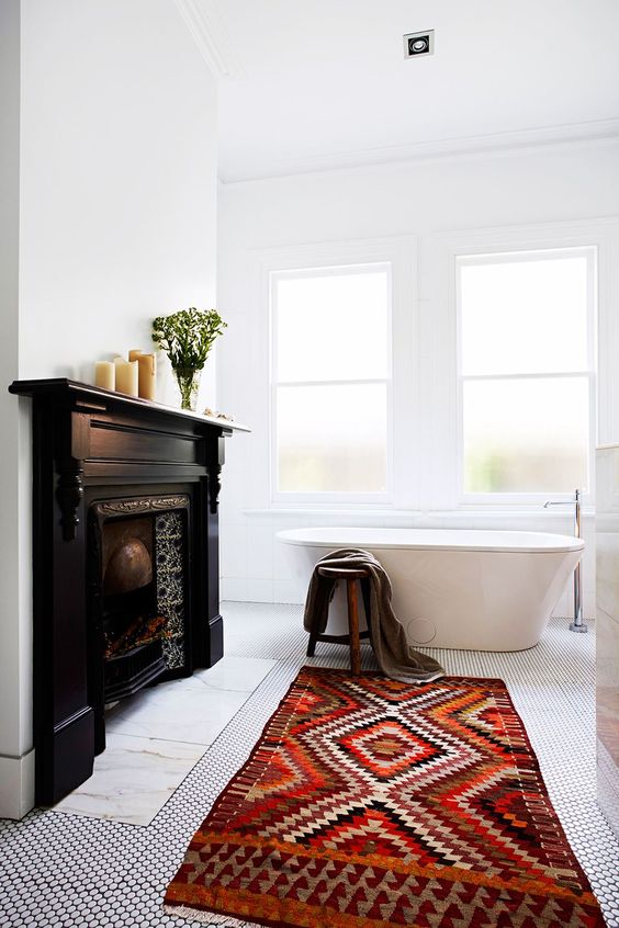 a contrasting bathroom with a vintage fireplace with a black mantel, a bright printed rug, an oval tub, greenery and candles