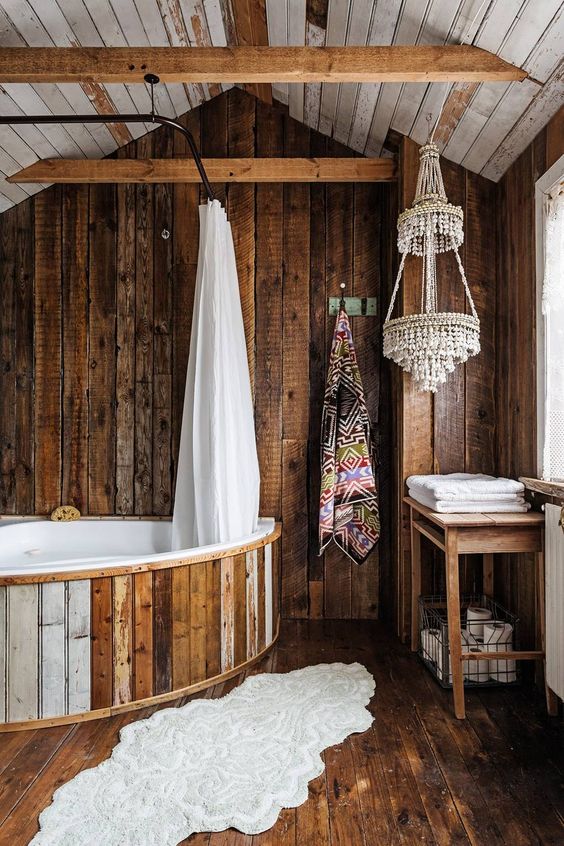 a creative bathroom clad with dark stained rough wood, with a bathtub clad with wood, beaded chandeliers and printed textiles