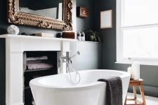 a fancy bathroom with black walls and a white planked floor, a fireplace used for storing towels, an oval tub and a mirror in a refined frame