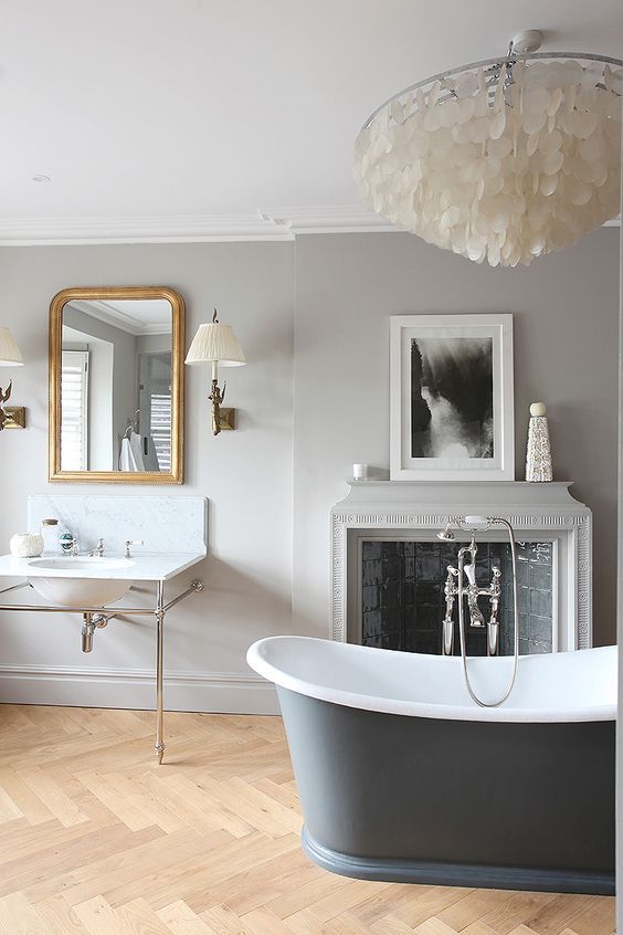 a neutral vintage bathroom with dove grey walls, a grey tub, a fireplace clad with tiles, a mother of pearl chandelier and a free standing tub