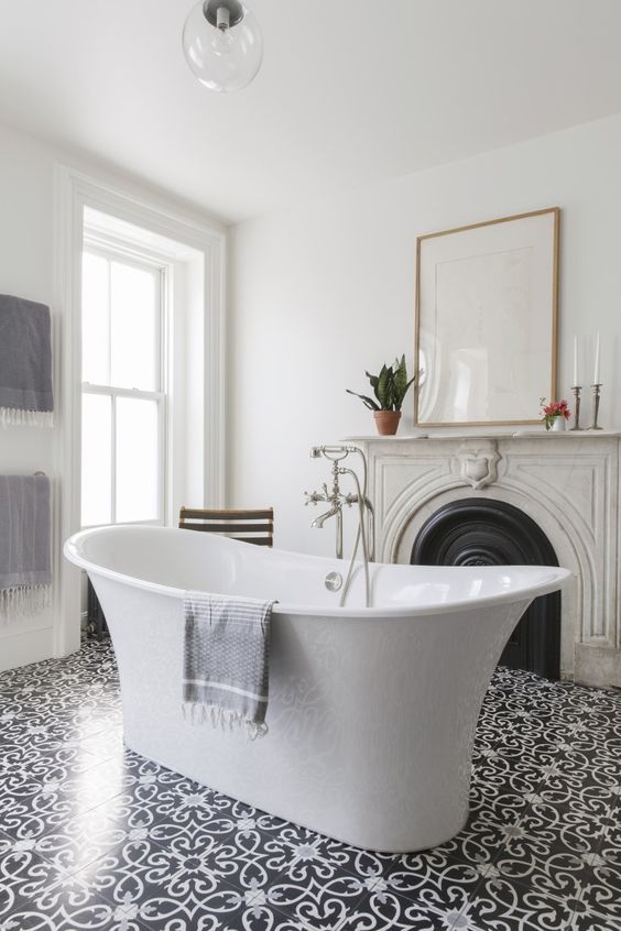 a refined and chic bathroom with a vintage fireplace with a stone mantel, a free-standing tub, some art and beautiful tiles on the floor