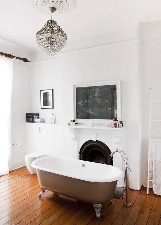 a refined bathroom with a rich-stained wooden floor, a non-working fireplace, lovely artworks, a crystal chandelier and a lovely bathtub