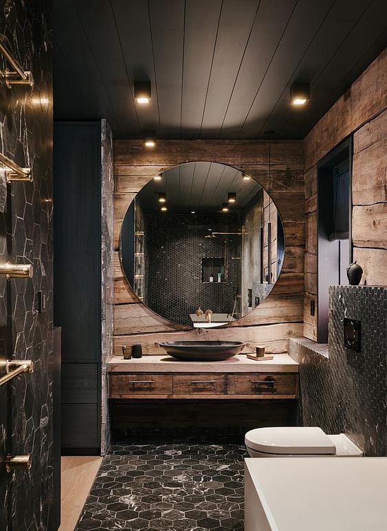 a refined moody bathroom clad with rough wood, with a black planked wooden ceiling, a round mirror, a tiled floor and matching walls