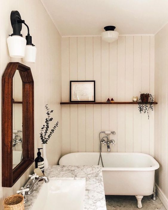 a small and cozy bathroom clad with white planks, a small tub, a vanity with a stone countertop, a mirror in a wooden frame