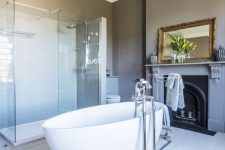 a stylish bathroom with grey walls, a vintage non-working fireplace, an oval tub, a shower enclosed in glass and a jute rug