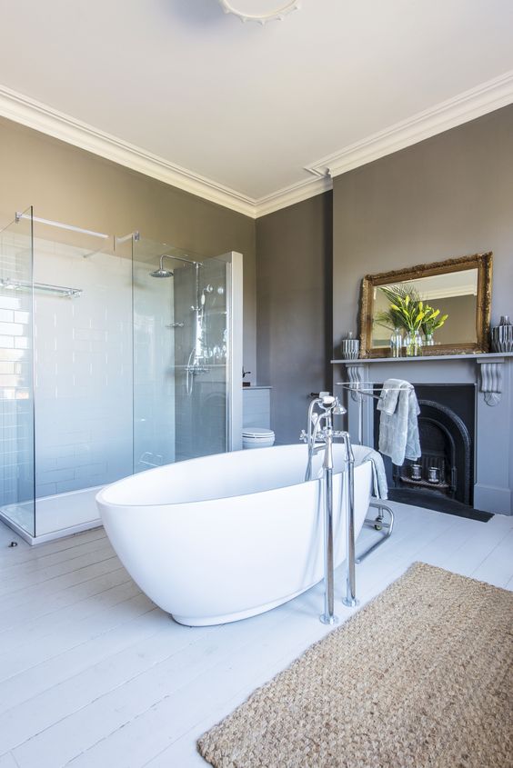 a stylish bathroom with grey walls, a vintage non working fireplace, an oval tub, a shower enclosed in glass and a jute rug
