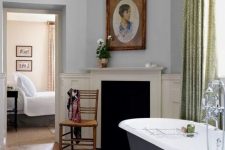 a vintage bathroom with grey walls, a faux fireplace, a navy clawfoot bathtub, green curtains, artwork and potted blooms