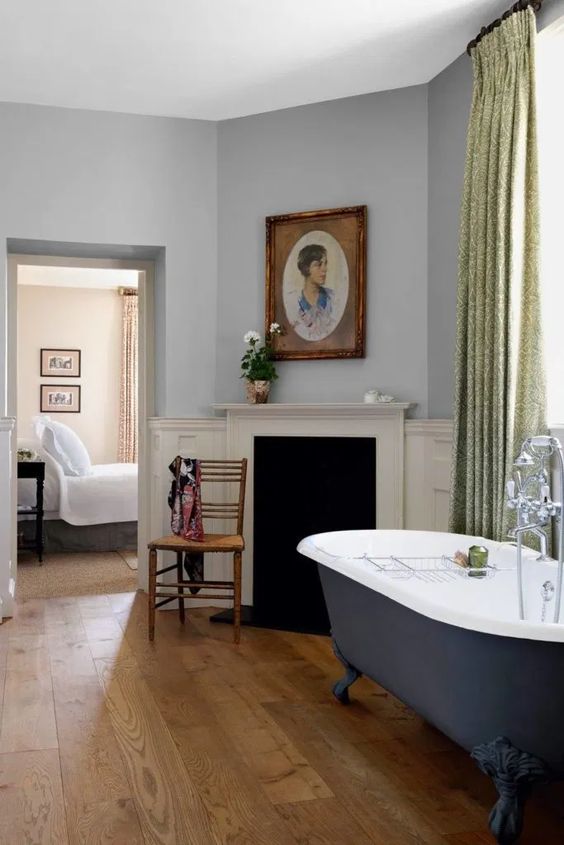 a vintage bathroom with grey walls, a faux fireplace, a navy clawfoot bathtub, green curtains, artwork and potted blooms