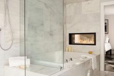 an exquisite white marble bathroom with a tub, a glass-enclosed shower space, a double-sided fireplace and some neutral textiles