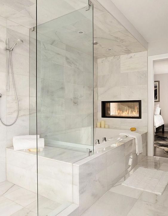 an exquisite white marble bathroom with a tub, a glass-enclosed shower space, a double-sided fireplace and some neutral textiles