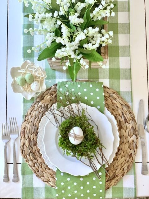 a green rustic Easter tablescape with a checked runner, a polka dot napkin, a woven placemat, lily of the valley centerpiece and a moss nest with an egg