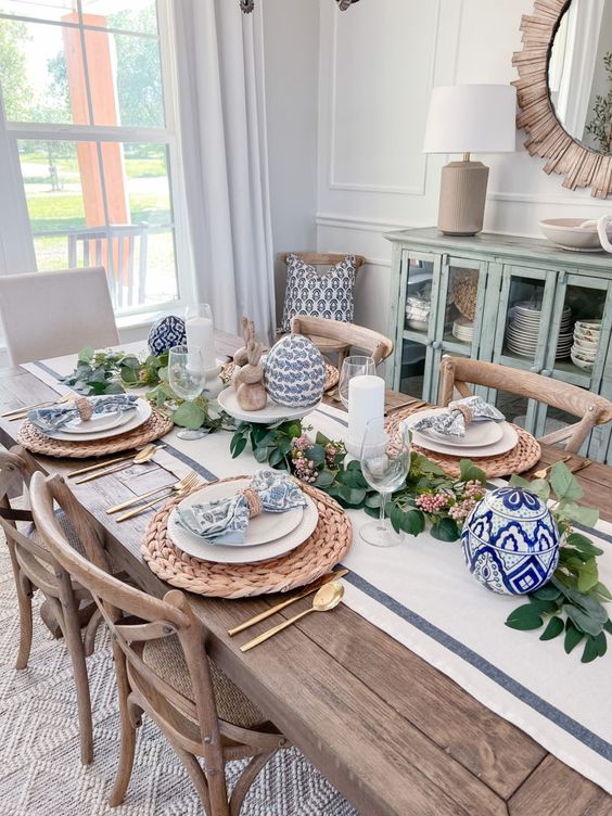 a lovely farmhouse Easter tablescape with a neutral runner, greenery, blue printed eggs and napkins, woven placemats and gold cutlery