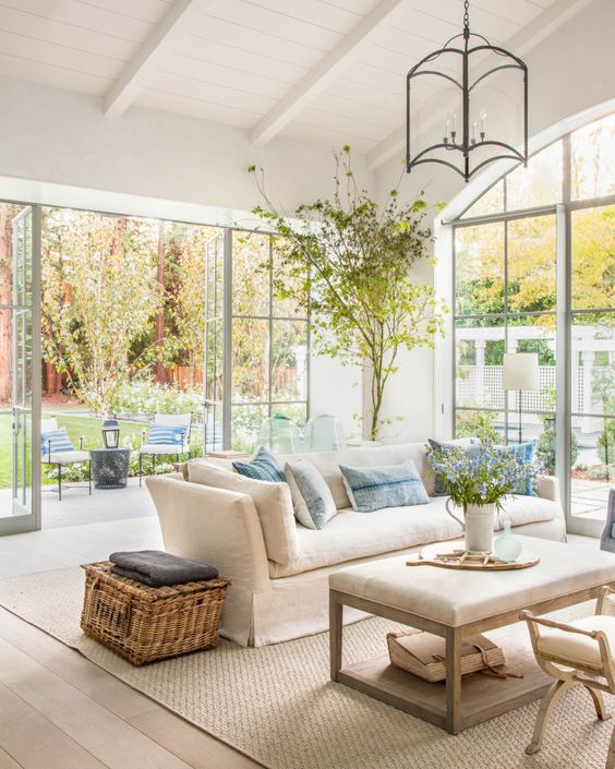 a neutral light-filled farmhouse living room with greenery, blooms and a tree in a pot is a chic space to be