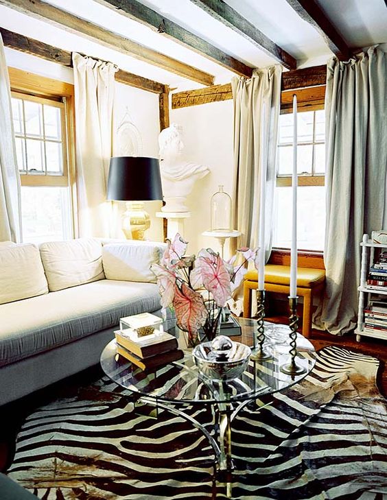 a beautiful living room with wooden beams, a white sofa, a leather side table, a shelving unit, a zebra print rug, a glass table with tall and thin candles