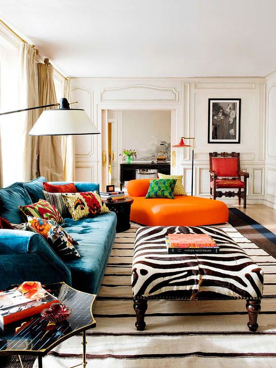 a beautiful maximalist living room with a blue sofa with colorful pillows, a zebra print ottoman, an orange daybed and cool floor lamps