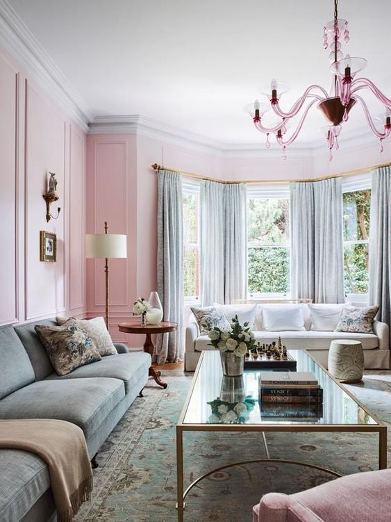 a beautiful pastel living room with pink walls, an aqua green sofa and matching curtains, a glass coffee table and a pretty pink chandelier