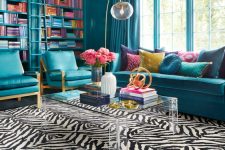 a bold and chic blue living room with bookcases, blue seating furniture, an acrylic table and a zebra print rug and touches of gold
