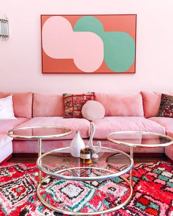 a bright and feminine living room with light pink walls, a pink sofa, a glass coffee table, a bold artwork and a colorful rug with prints