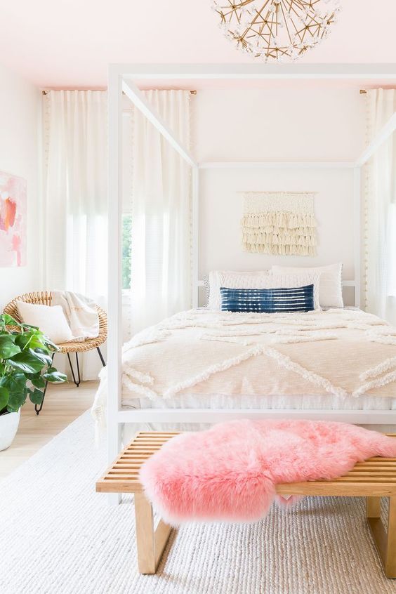 a bright feminine bedroom with a cnaopy bed, a wooden bench and a woven chair, a statement plant and a floral chandelier