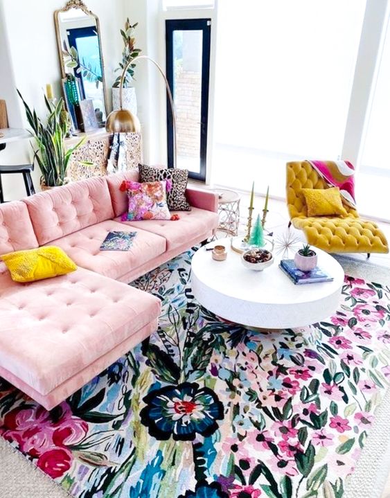 a bright girlish living room with a colorful floral print rug, a pink sofa, a mustard lounger, colorful pillows and potted greenery