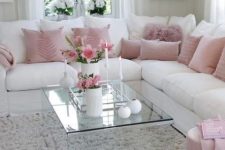a chic and elegant living room with a white corner sofa, pink pillows and a pouf, a glass coffee table and a crystal chandelier
