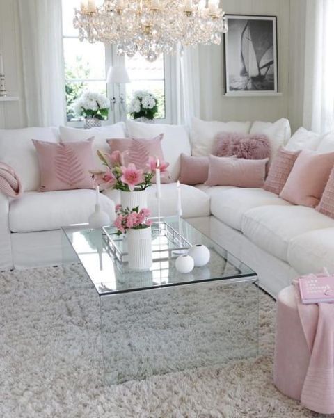 a chic and elegant living room with a white corner sofa, pink pillows and a pouf, a glass coffee table and a crystal chandelier